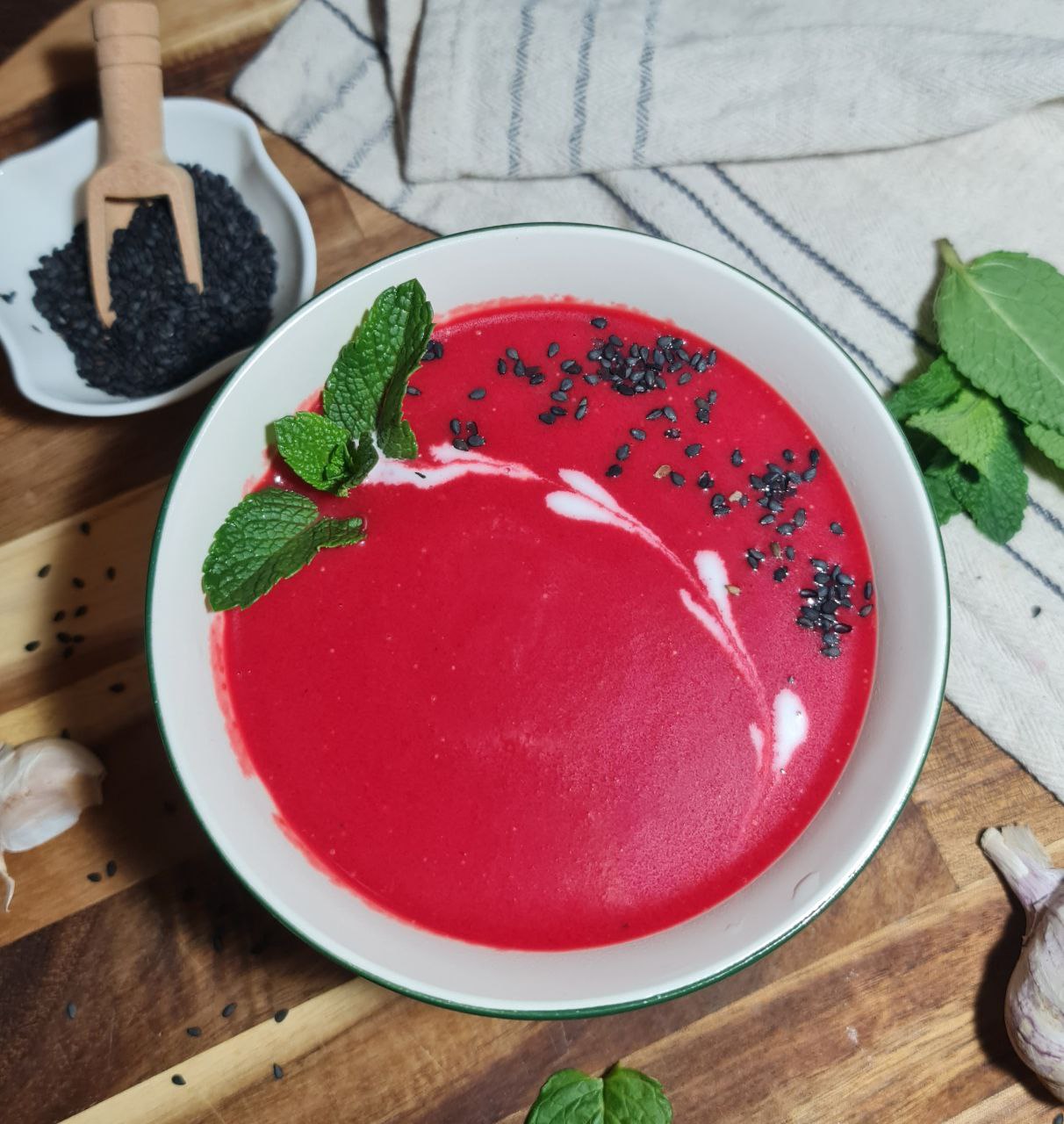 A bowl of creamy vegan beet soup garnished with fresh mint leaves and black sesame seeds, placed on a wooden surface with garlic cloves, mint leaves, and a small dish of black sesame seeds nearby.