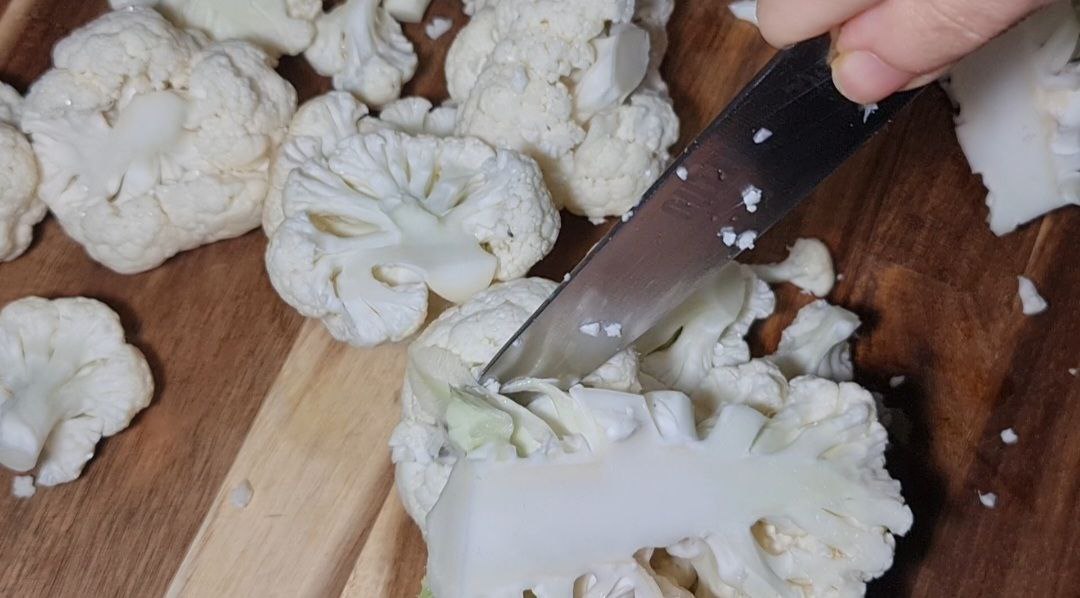 Close-up of a knife cutting cauliflower florets on a wooden cutting board.