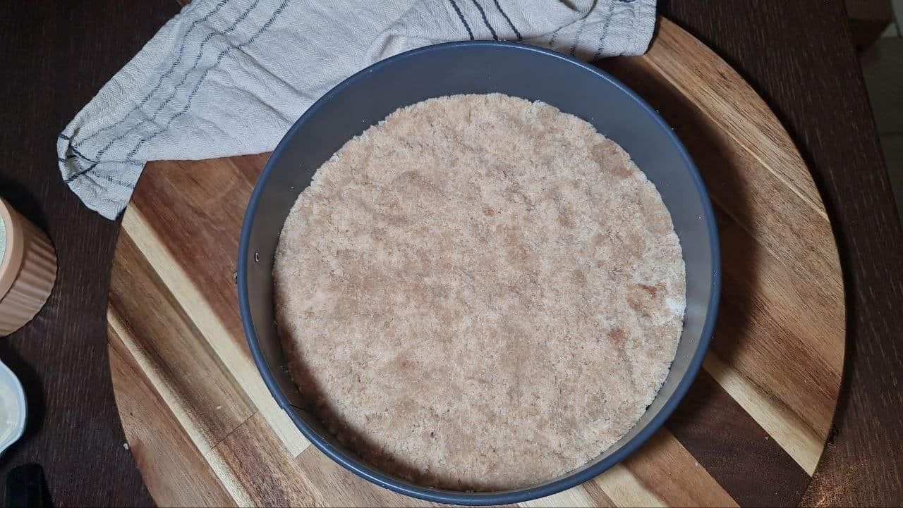 A close-up of a prepared crust in a round baking pan, set on a wooden cutting board with a cloth in the background, ready for a guilt-free cake recipe.