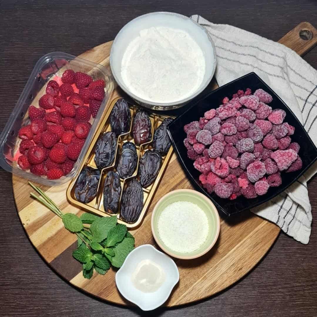 - Ingredients for a guilt-free cake recipe including fresh raspberries, dates, frozen raspberries, mint leaves, and various powders on a wooden board.
- Caption: A selection 