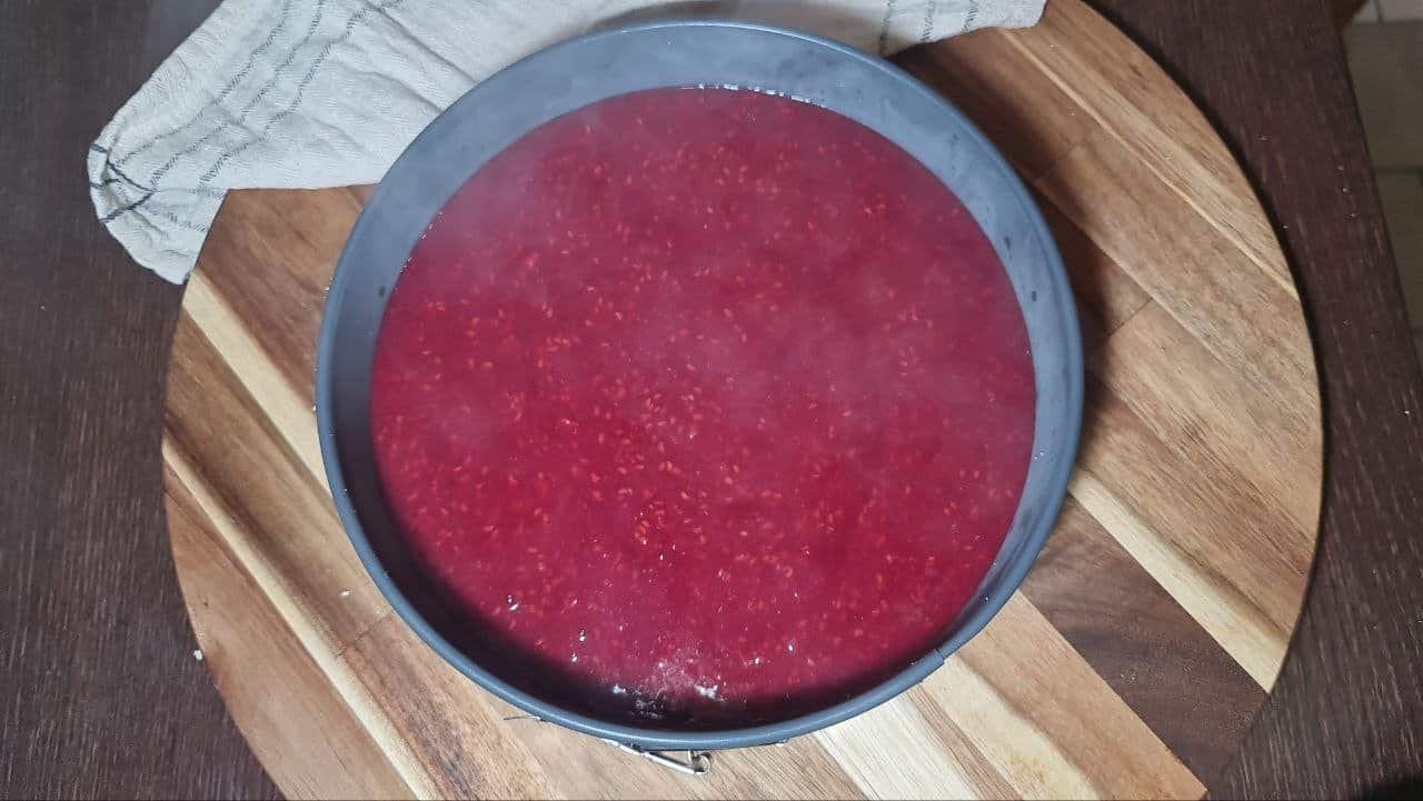 A round cake pan filled with a vibrant red raspberry mixture, placed on a wooden cutting board.