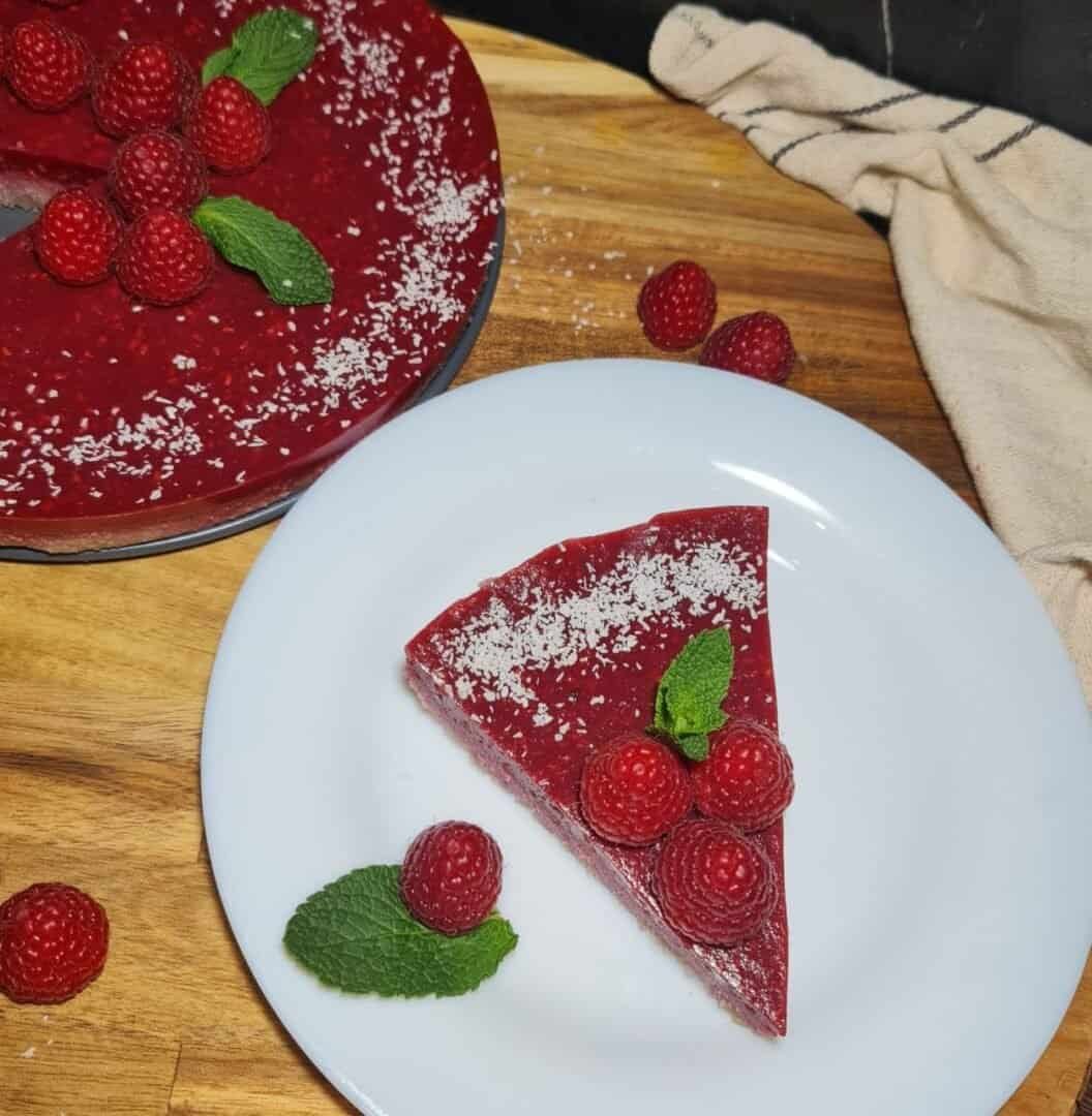 A slice of guilt-free raspberry cake garnished with fresh raspberries and mint leaves, served on a white plate with the remaining cake in the background.