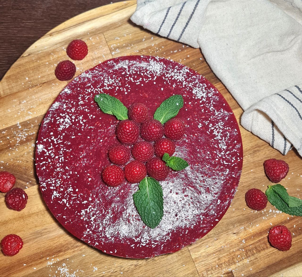 A vibrant red guilt-free raspberry cake topped with fresh raspberries and mint leaves, sprinkled with coconut flakes, placed on a wooden board with a striped cloth napkin.