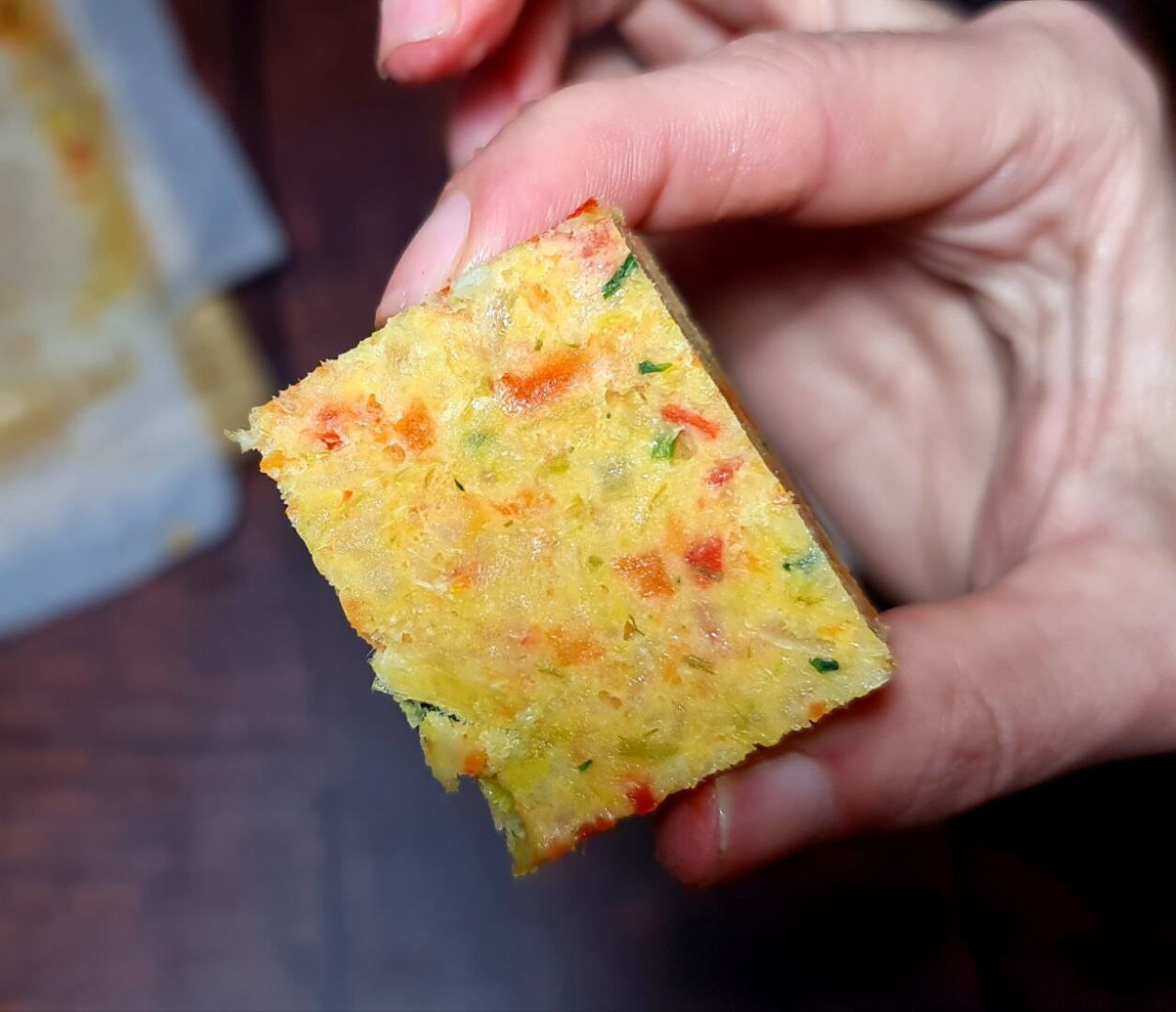 Close-up of a homemade vegetable broth cube held between fingers, showing a colorful mix of finely chopped vegetables.