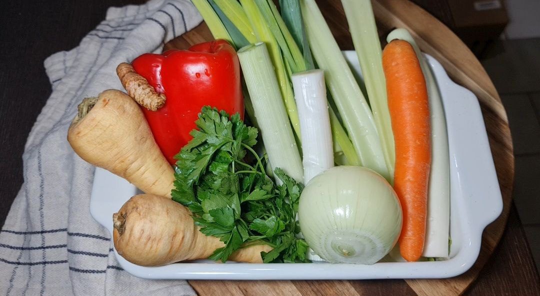 Fresh vegetables including parsnips, red bell pepper, leeks, carrot, onion, and parsley in a white dish on a wooden board.