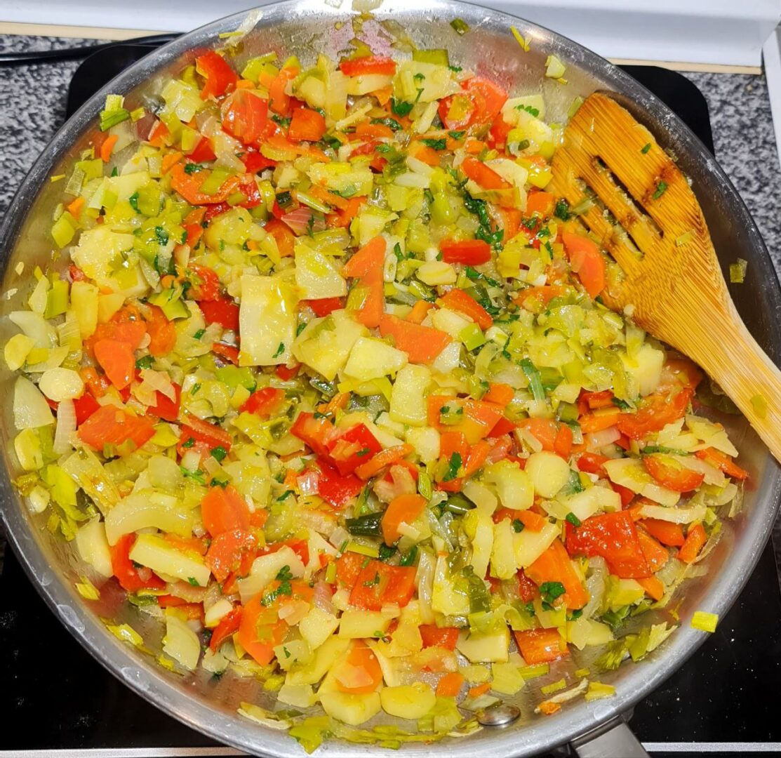 A pan filled with a colorful mixture of chopped vegetables including carrots, red bell peppers, onions, and herbs, being sautéed with a wooden spatula.