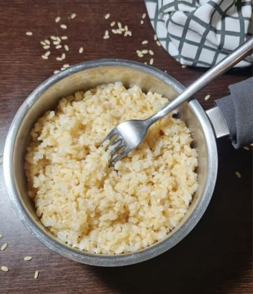 A pot of cooked brown rice with a fork, placed on a wooden surface with scattered rice grains and a checkered cloth in the background.