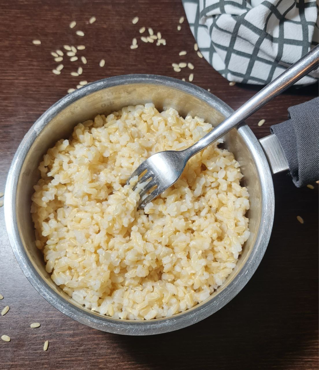 A pot of cooked brown rice with a fork, placed on a wooden surface with scattered rice grains and a checkered cloth in the background.