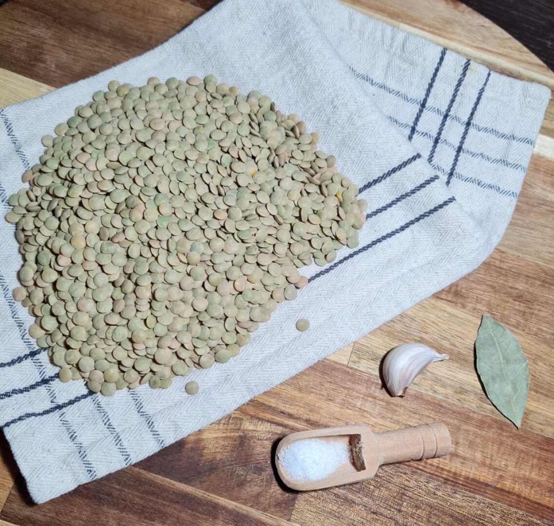 A pile of green lentils on a cloth napkin with a garlic clove, bay leaf, and a wooden scoop of salt on a wooden surface.