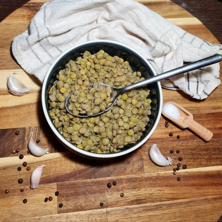 A bowl of cooked lentils with a spoon, surrounded by garlic cloves, black peppercorns, and a small wooden scoop of salt on a wooden surface.