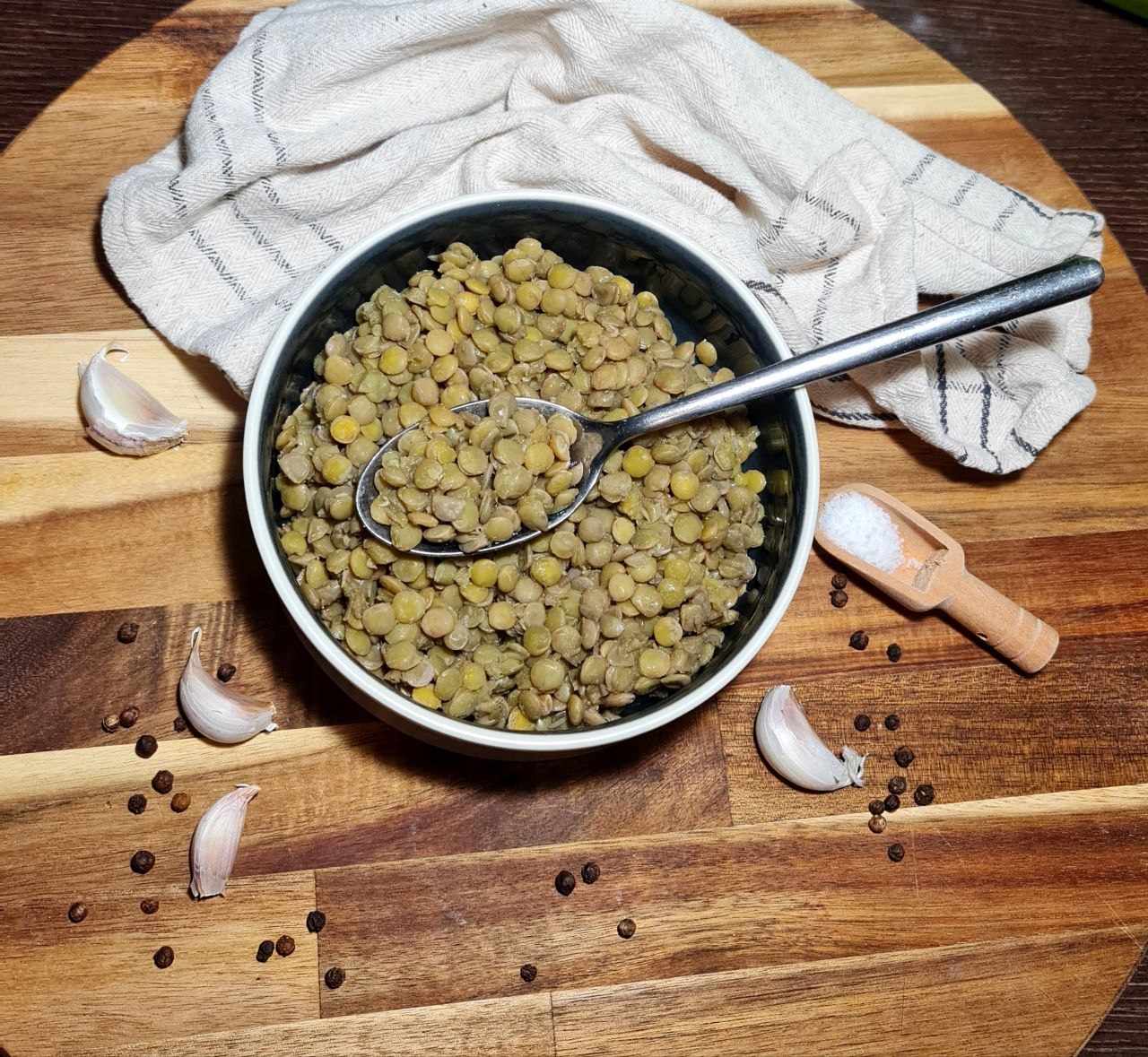 A bowl of cooked lentils with a spoon, surrounded by garlic cloves, black peppercorns, and a small wooden scoop of salt on a wooden surface.