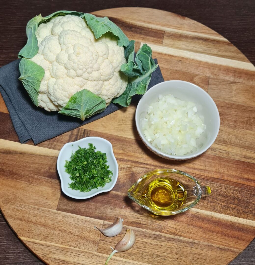 Ingredients for making cauliflower rice including a whole cauliflower, chopped onions, chopped parsley, olive oil, and garlic cloves on a wooden board.