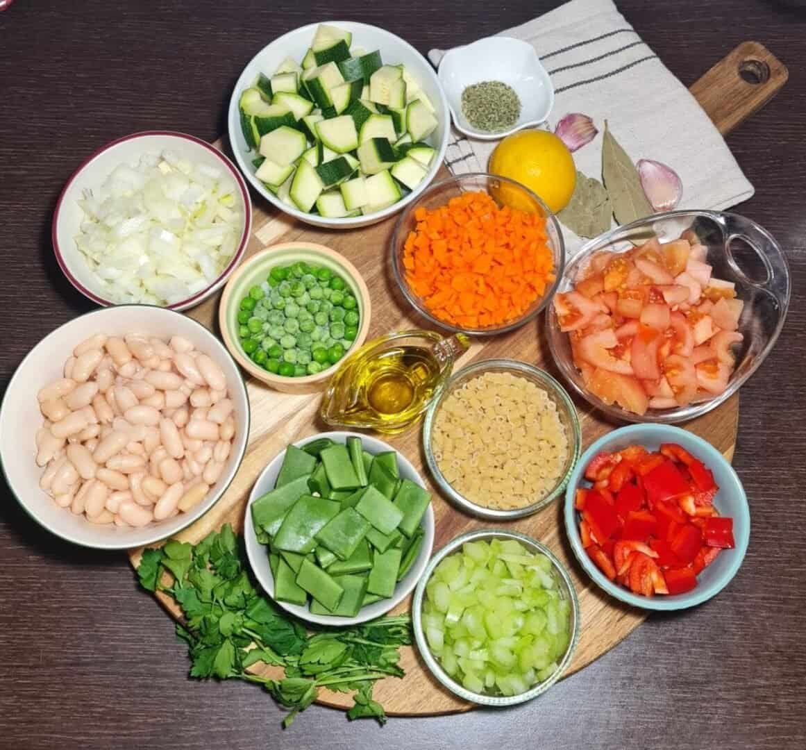  Ingredients for minestrone soup including chopped zucchini, diced onions, white beans, frozen peas, olive oil, diced carrots, chopped tomatoes, small pasta, red bell peppers, celery, green beans, fresh parsley, garlic, lemon, bay leaves, and dried herbs.