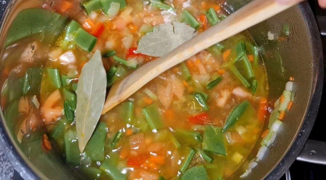 A pot of minestrone soup being stirred with a wooden spoon, containing green beans, diced tomatoes, carrots, onions, and bay leaves.