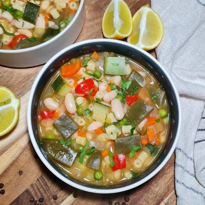 A bowl of colorful minestrone soup filled with vegetables, beans, and pasta, garnished with fresh herbs, with lemon wedges on the side.