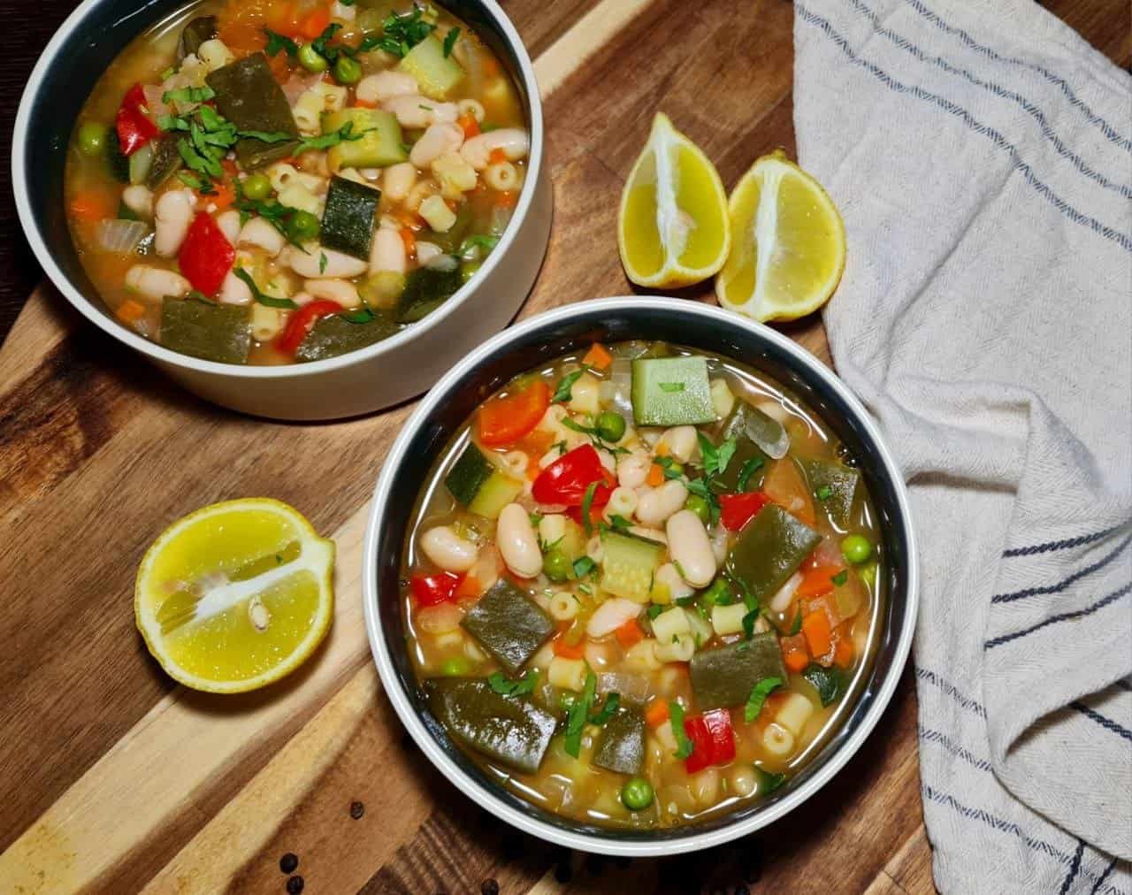 Two bowls of colorful minestrone soup with vegetables and pasta, garnished with fresh herbs, served with lemon wedges on a wooden board.