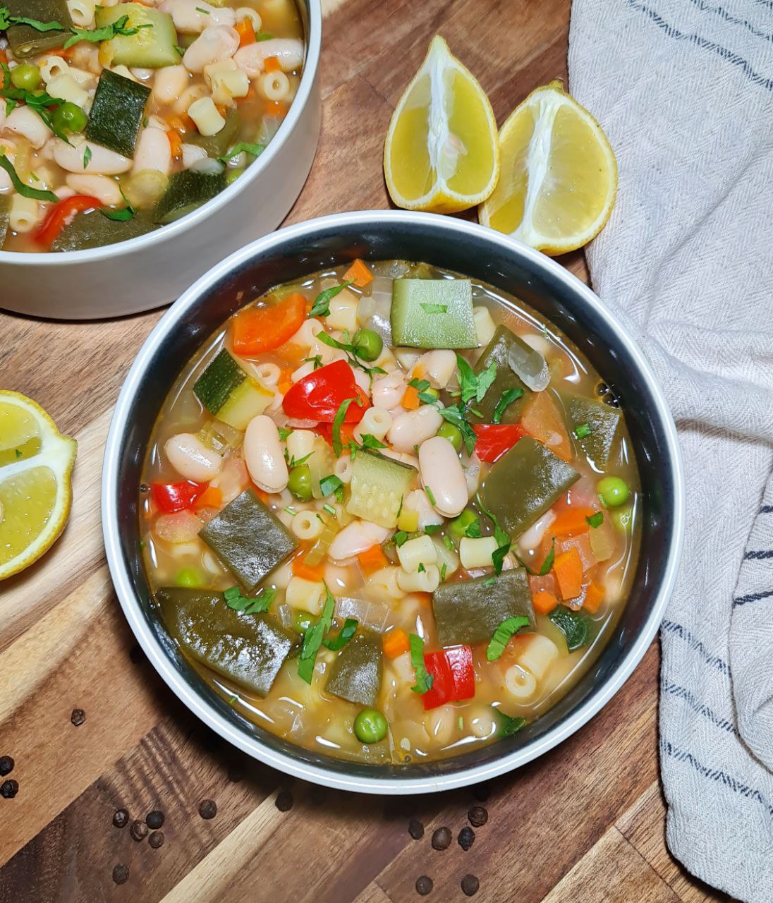 A bowl of colorful minestrone soup filled with vegetables, beans, and pasta, garnished with fresh herbs, with lemon wedges on the side.