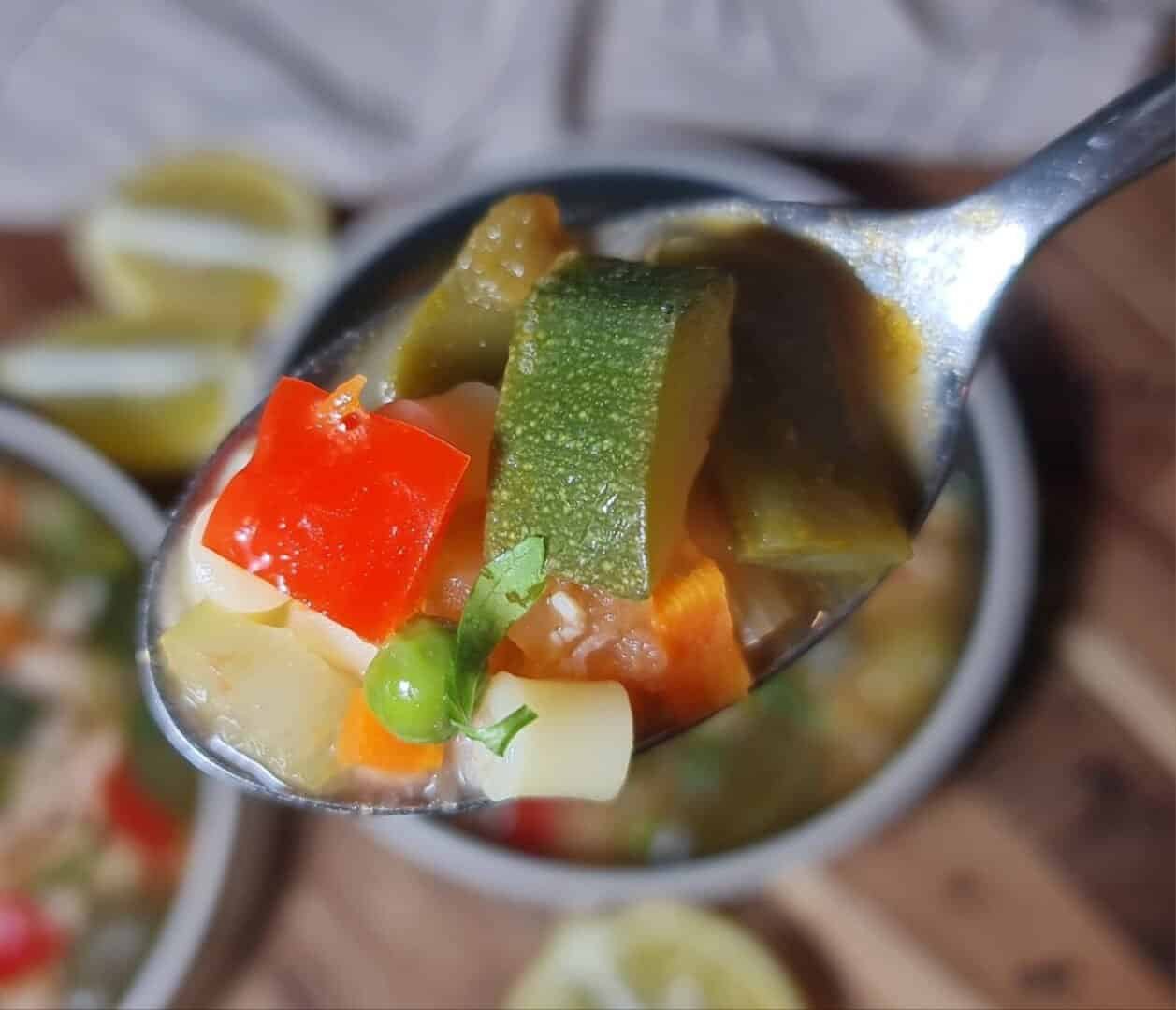 Close-up of a spoonful of minestrone soup with various vegetables including zucchini, red bell pepper, carrot, pea, and pasta.