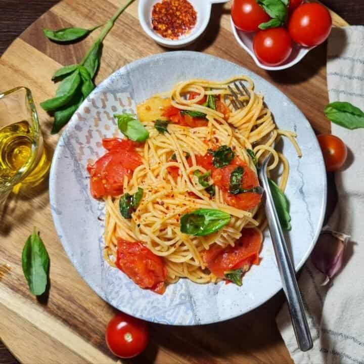 A bowl of spaghetti pasta with cherry tomatoes, fresh basil, and a sprinkle of red pepper flakes, surrounded by fresh ingredients on a wooden board.