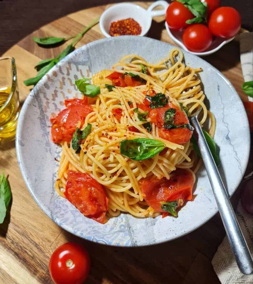 A plate of spaghetti pasta garnished with cherry tomatoes, fresh basil leaves, and red pepper flakes, accompanied by a small bowl of red pepper flakes and fresh cherry tomatoes on a wooden table.