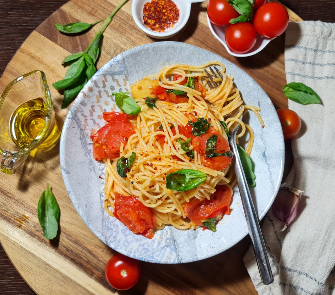 A bowl of spaghetti pasta with cherry tomatoes, fresh basil, and a sprinkle of red pepper flakes, surrounded by fresh ingredients on a wooden board.