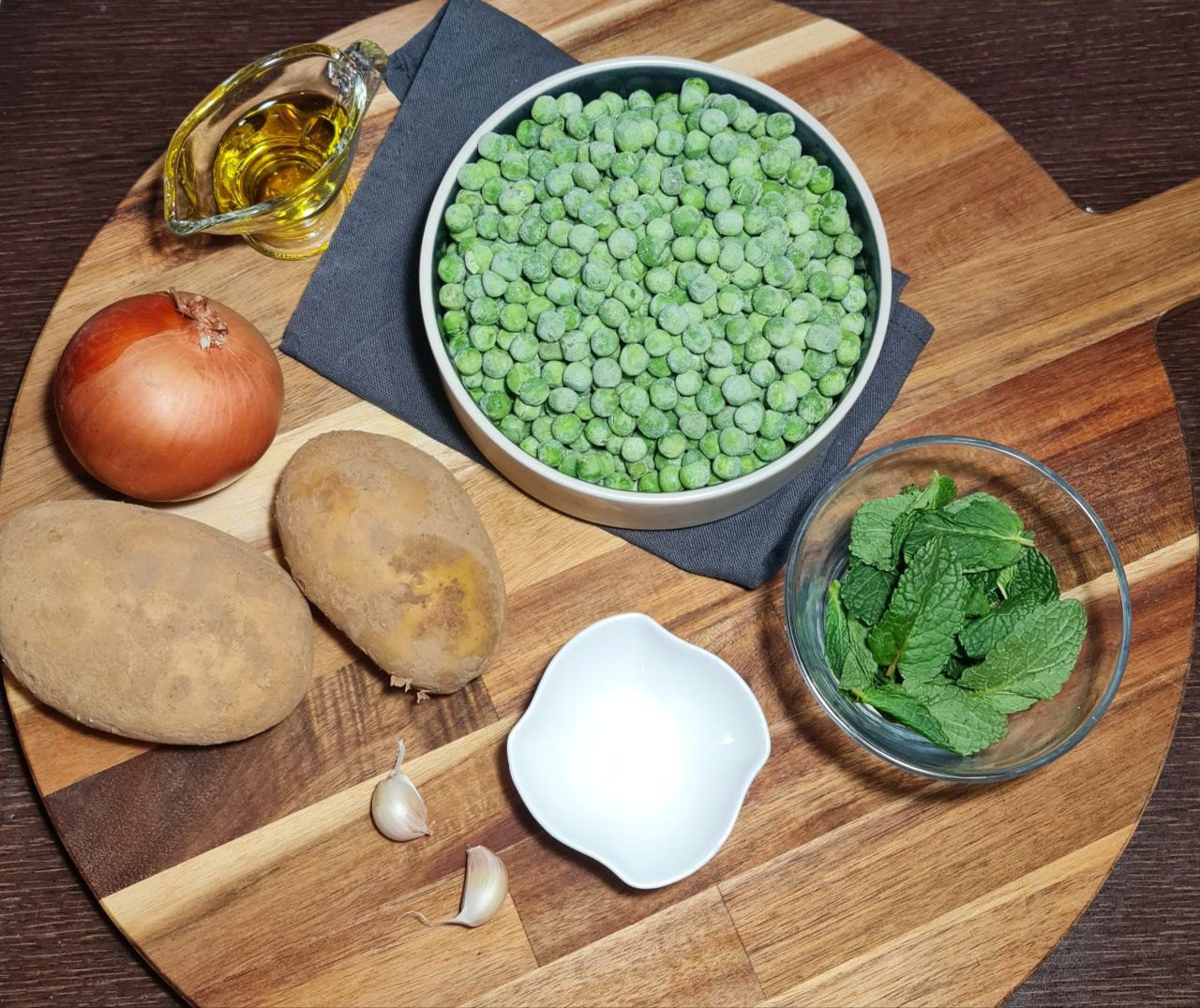 Ingredients for pea and mint soup including frozen peas, fresh mint leaves, potatoes, onion, garlic, and olive oil on a wooden cutting board.