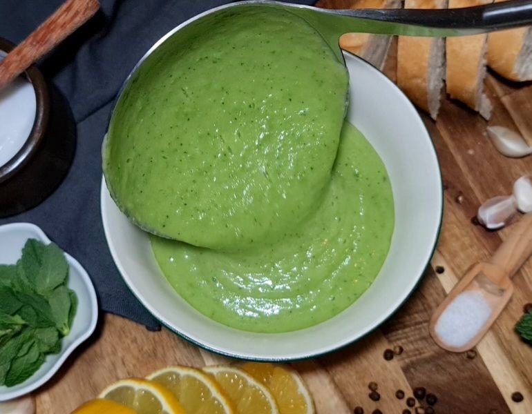 A ladle pouring creamy green pea and mint soup into a white bowl, surrounded by fresh ingredients like mint leaves, lemon slices, garlic, and bread.