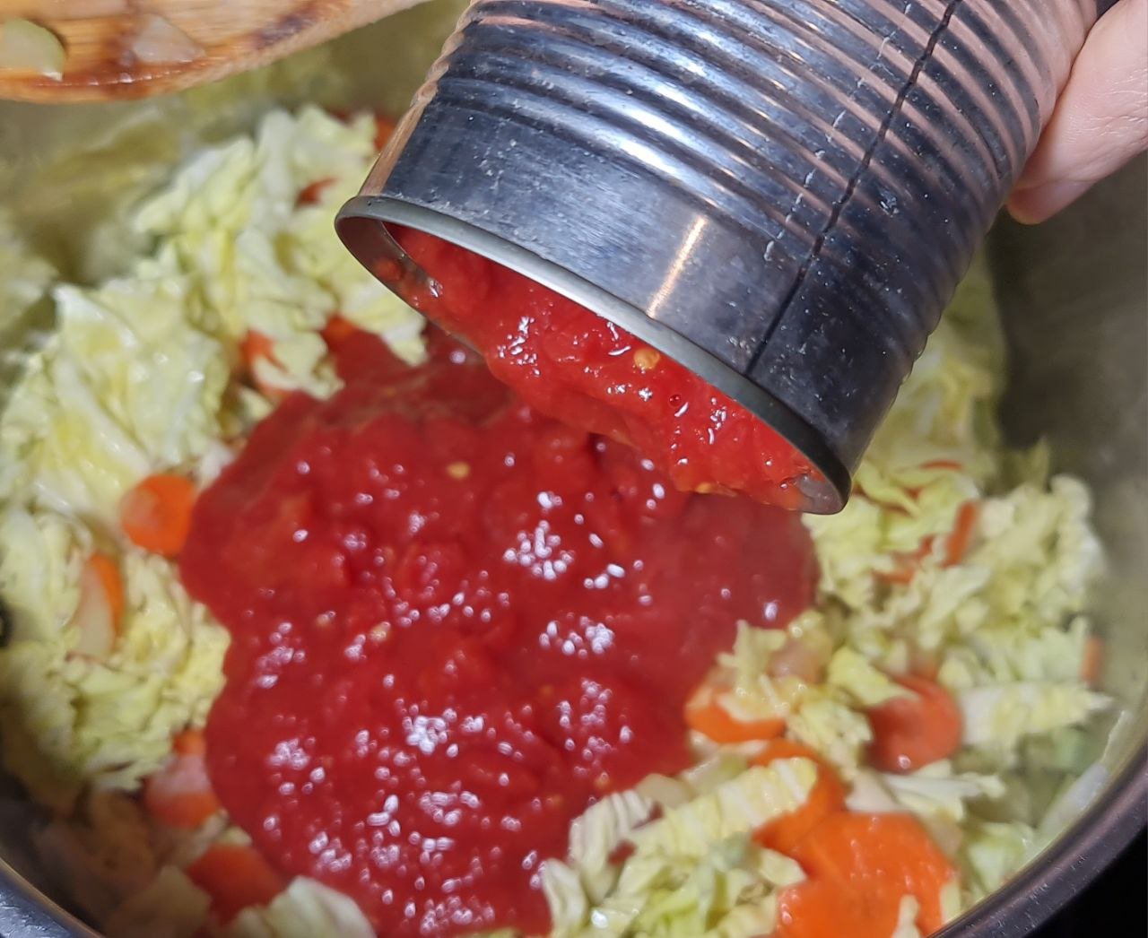 Pouring tomato sauce from a can into a pot of cabbage and carrots for cabbage soup.