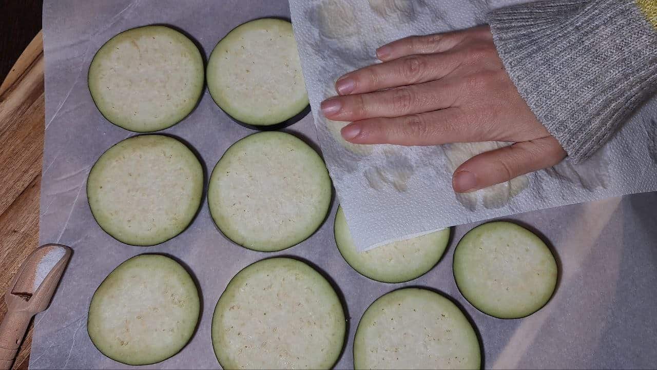 Hand pressing paper towel on eggplant slices to remove moisture before sautéing.