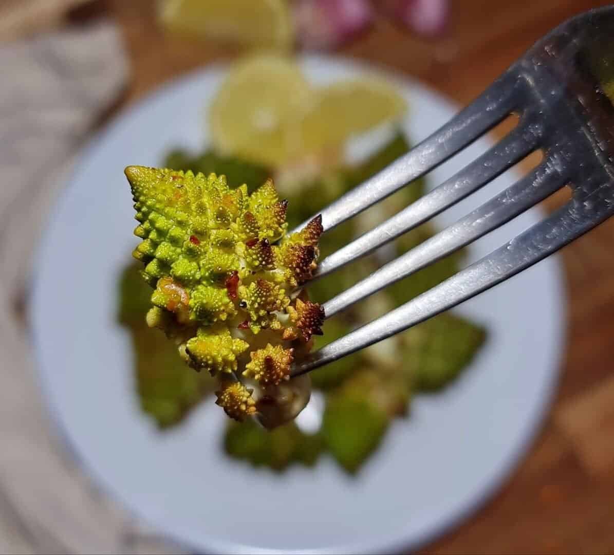 Close-up of a fork holding a piece of roasted Romanesco with a plate of Romanesco and lemon slices in the background.