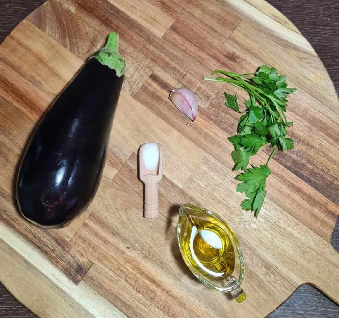Ingredients for sautéed eggplant recipe including an eggplant, garlic clove, fresh parsley, salt, and olive oil on a wooden cutting board.