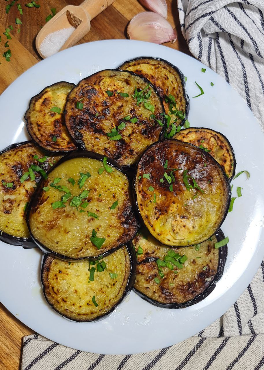 A plate of sauteed eggplant slices garnished with chopped herbs, placed on a wooden surface with a small wooden scoop of salt and garlic cloves in the background.