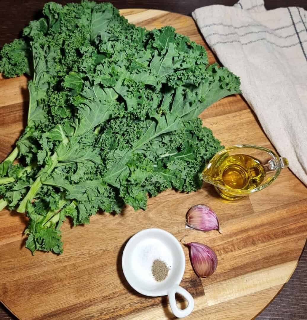 Fresh kale leaves, olive oil, garlic cloves, salt, and pepper on a wooden cutting board with a kitchen towel.