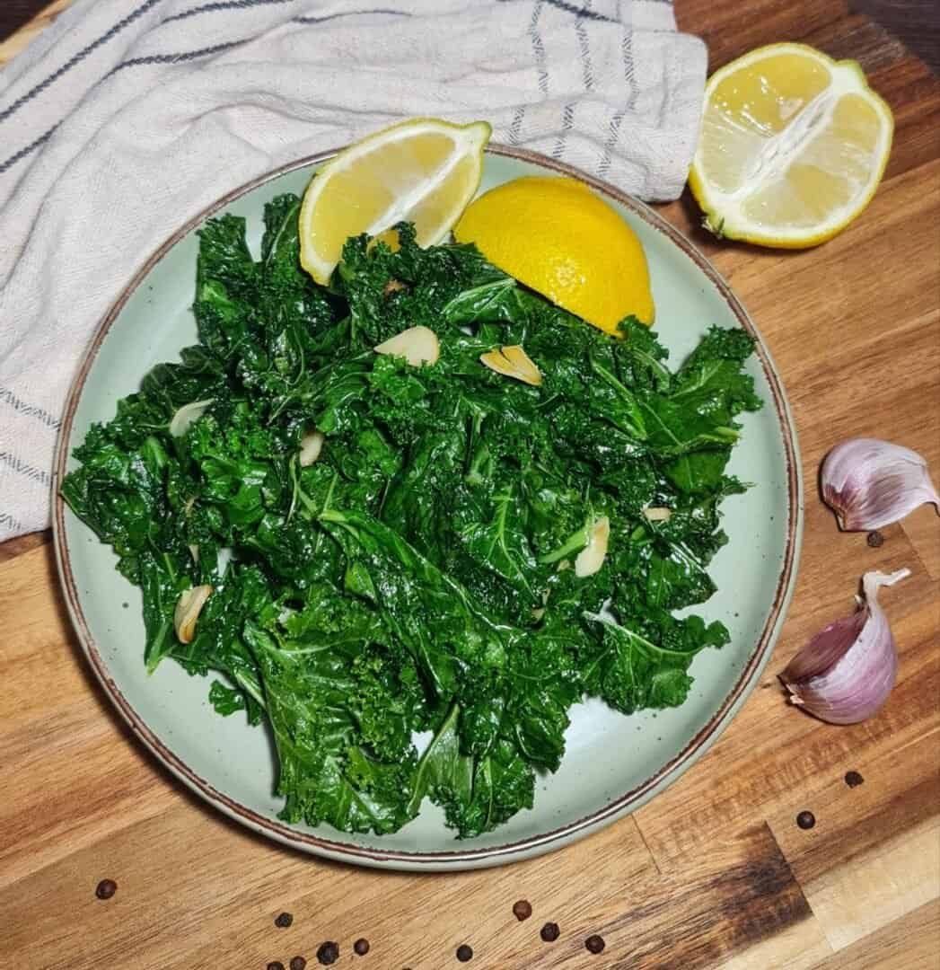  A plate of sauteed kale garnished with garlic slices and lemon wedges, placed on a wooden cutting board with garlic cloves and a striped cloth in the background.