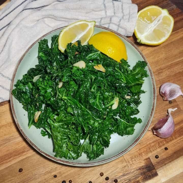 A plate of sauteed kale garnished with garlic slices and lemon wedges, placed on a wooden cutting board with garlic cloves and a striped cloth in the background.