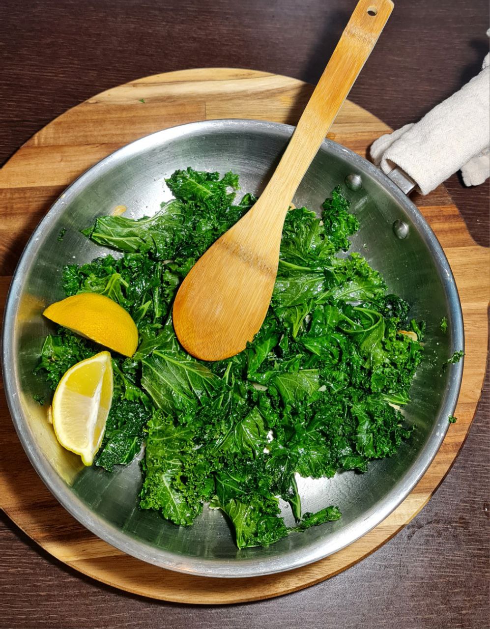 A pan of freshly sauteed kale with lemon wedges and a wooden spatula.