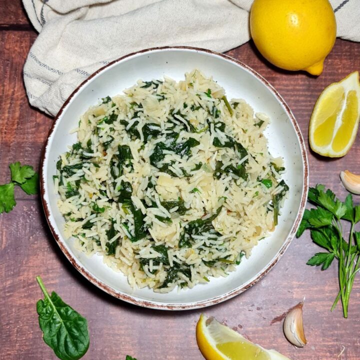 A bowl of spinach rice garnished with fresh parsley, lemon wedges, and garlic cloves on a wooden table.