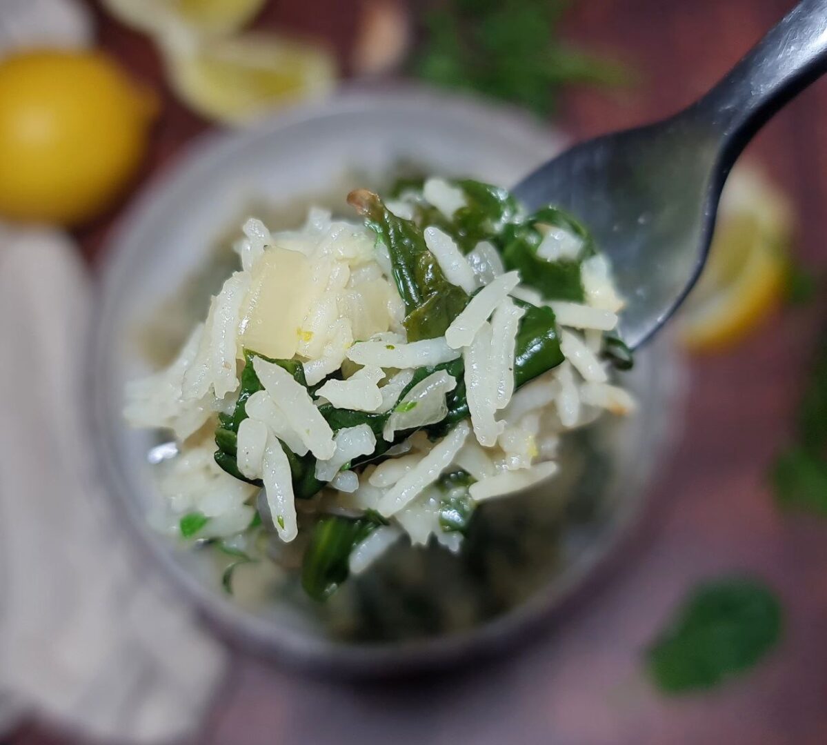 Close-up of a forkful of spinach rice with visible spinach leaves and rice grains, with a blurred background of lemon slices and fresh herbs.