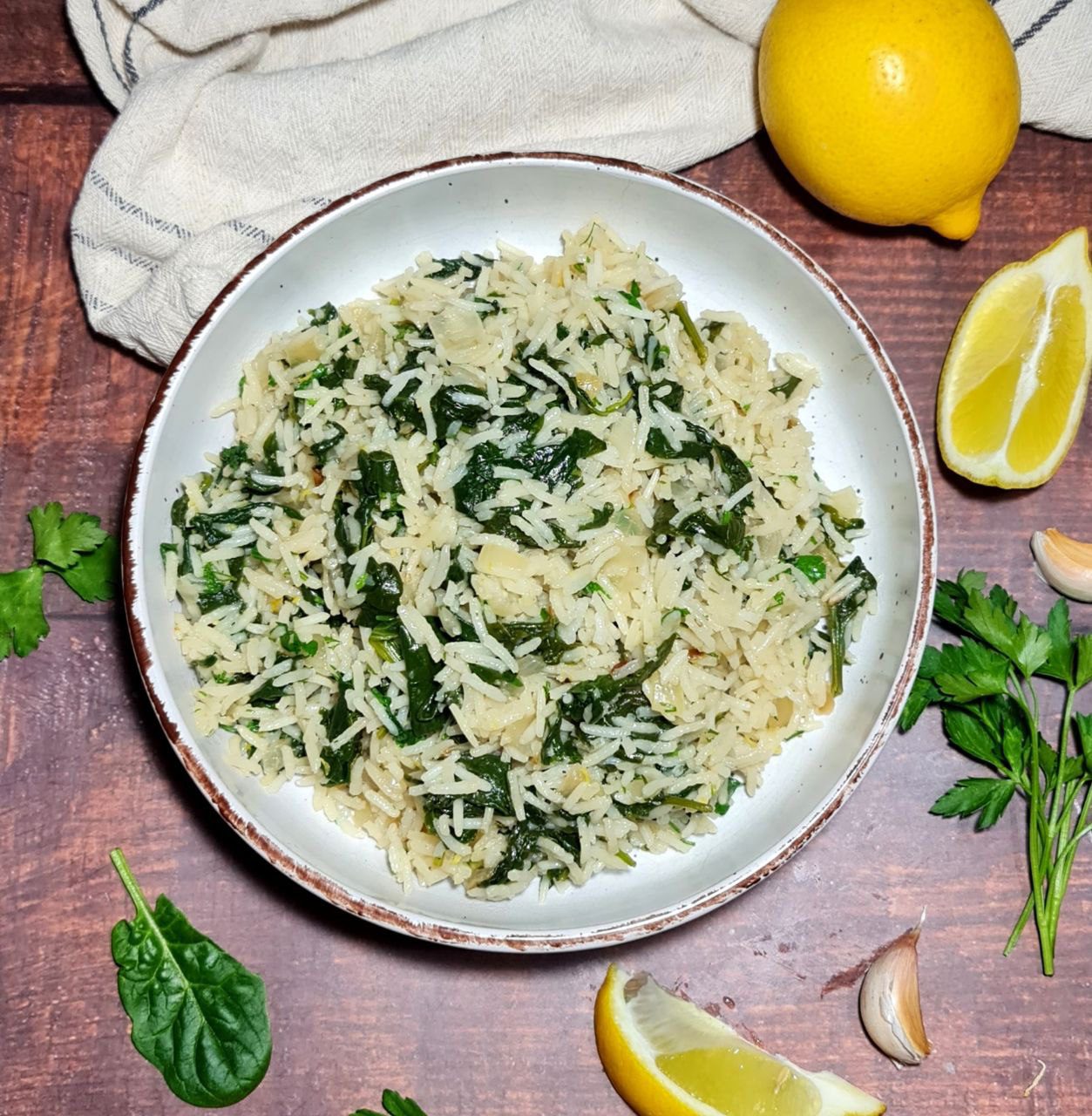 A bowl of spinach rice garnished with fresh parsley, lemon wedges, and garlic cloves on a wooden table.