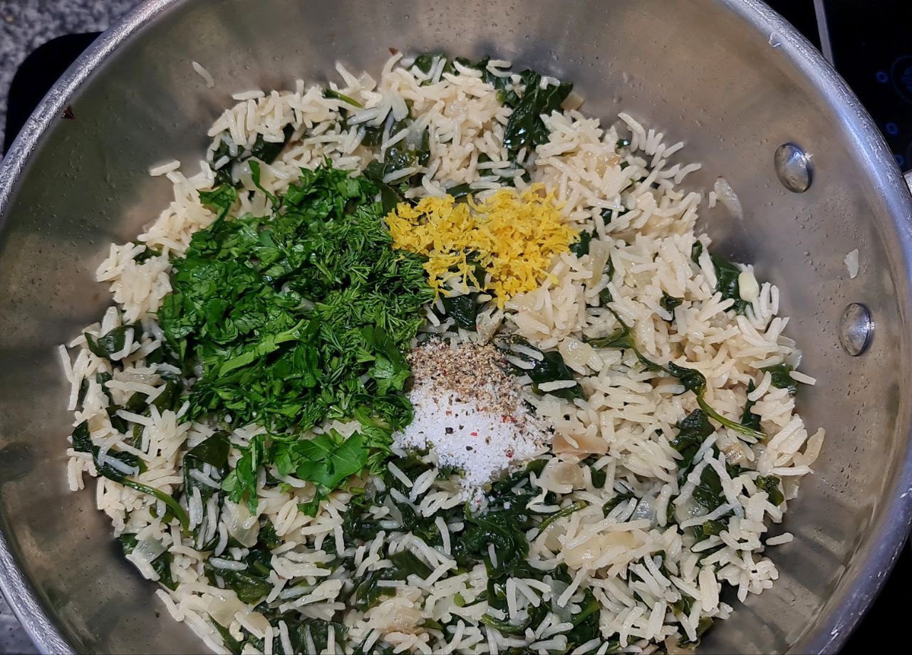 A close-up view of a pot containing cooked rice mixed with spinach, fresh herbs, lemon zest, salt, and pepper.