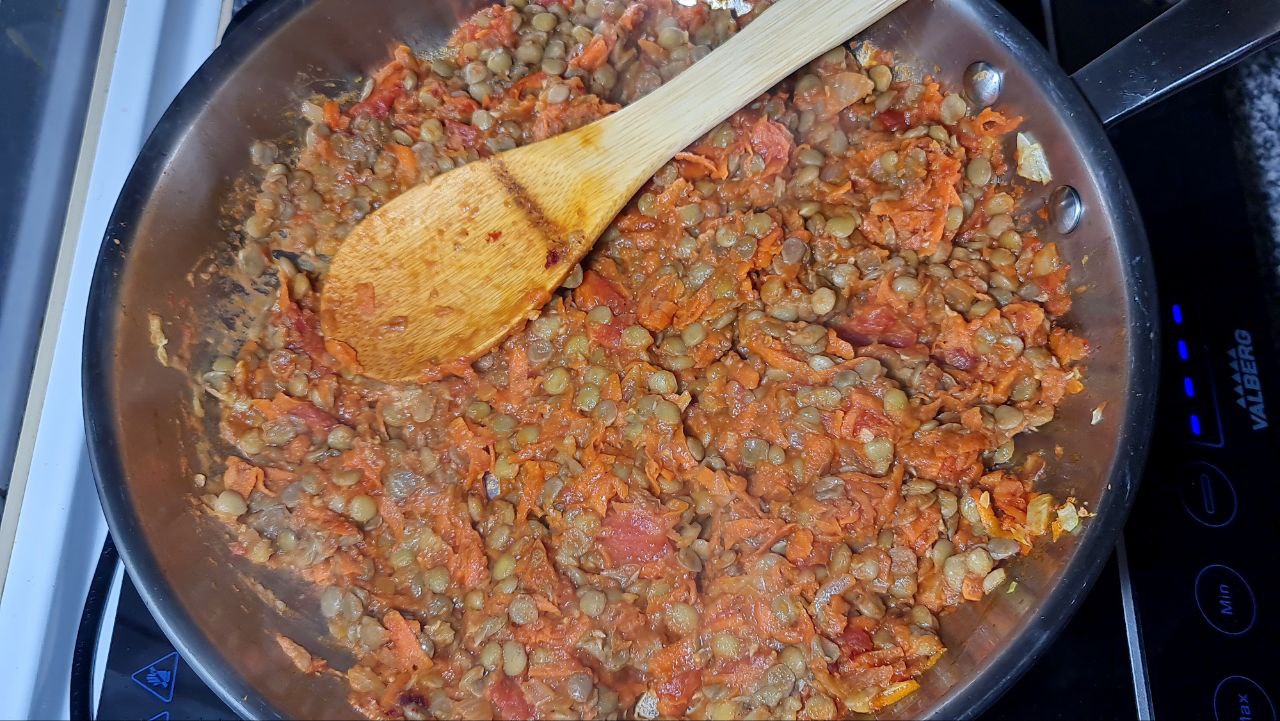 A pan filled with a mixture of lentils, carrots, tomatoes, and spices being stirred with a wooden spoon.