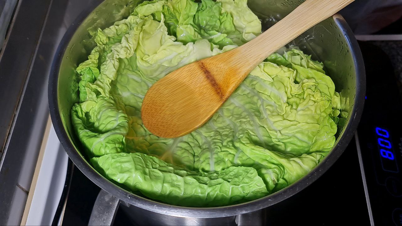 Fresh green cabbage leaves being blanched in a pot of boiling water with a wooden spoon.