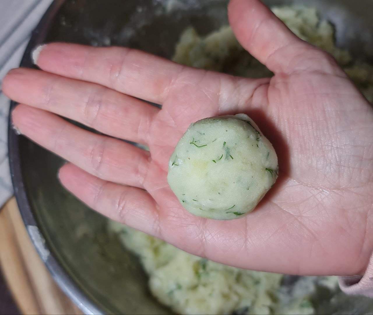 A hand holding a vegan mashed potato ball with dill, ready to be cooked.