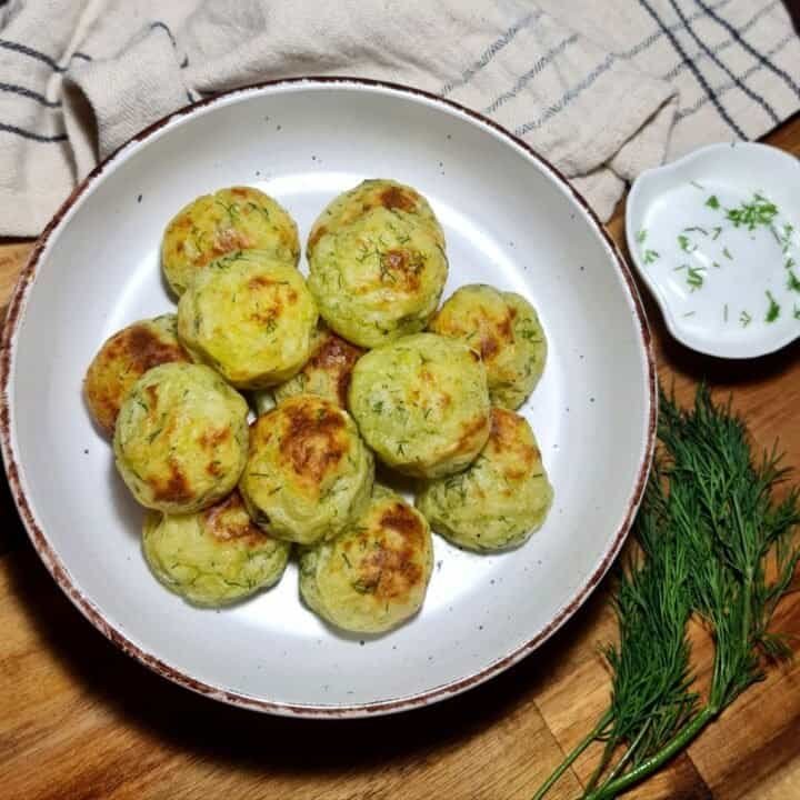 A plate of golden-brown vegan mashed potato balls garnished with fresh dill, served on a white dish with a side of chopped herbs.