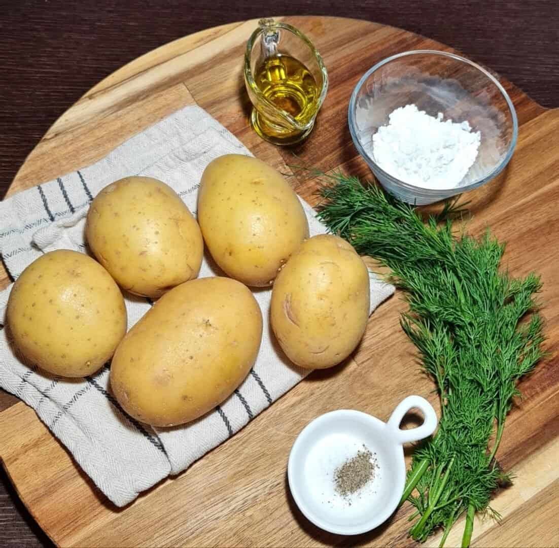 Ingredients for vegan mashed potato balls including potatoes, olive oil, cornstarch, fresh dill, salt, and pepper on a wooden board.