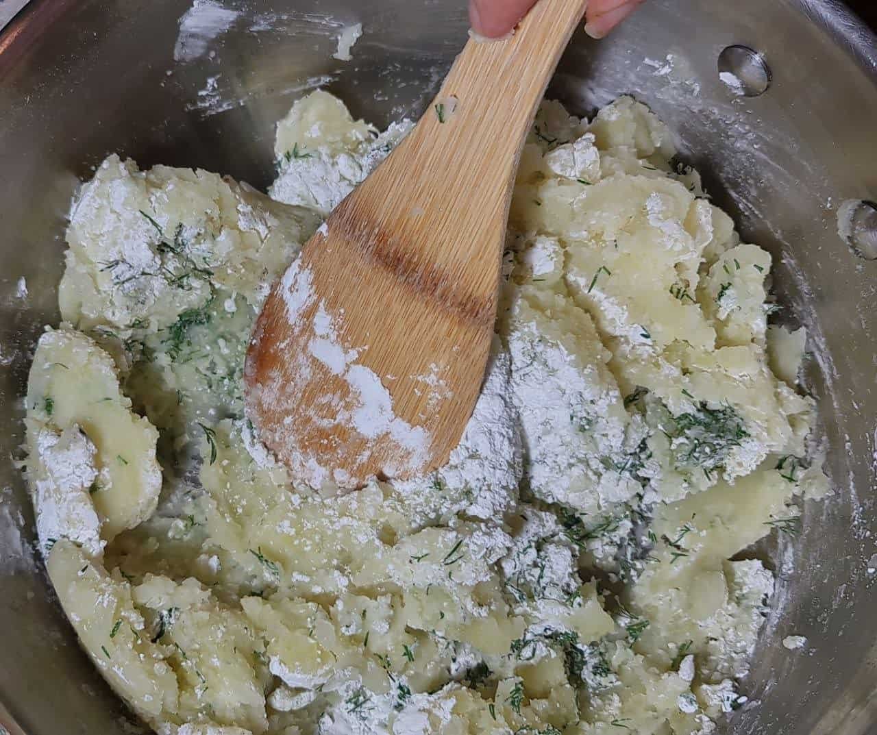 Mixing vegan mashed potato mixture with flour and herbs in a metal bowl using a wooden spoon.