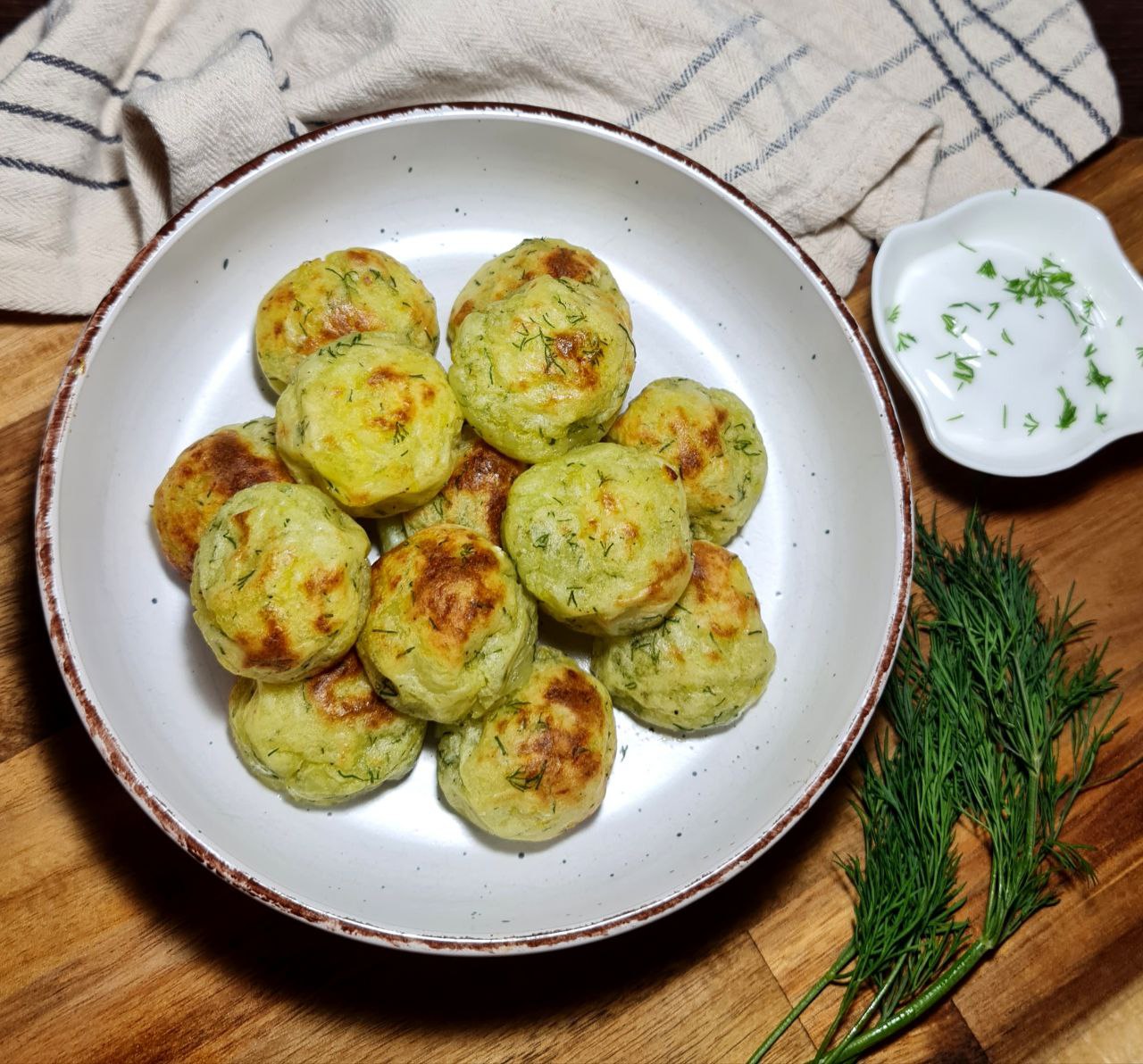 A plate of golden-brown vegan mashed potato balls garnished with fresh dill, served on a white dish with a side of chopped herbs.