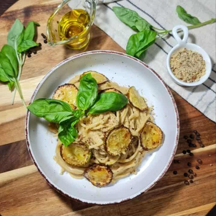 A bowl of vegan spaghetti alla Nerano garnished with fresh basil leaves and roasted zucchini slices, surrounded by ingredients including olive oil, ground nuts, and fresh basil on a wooden surface.