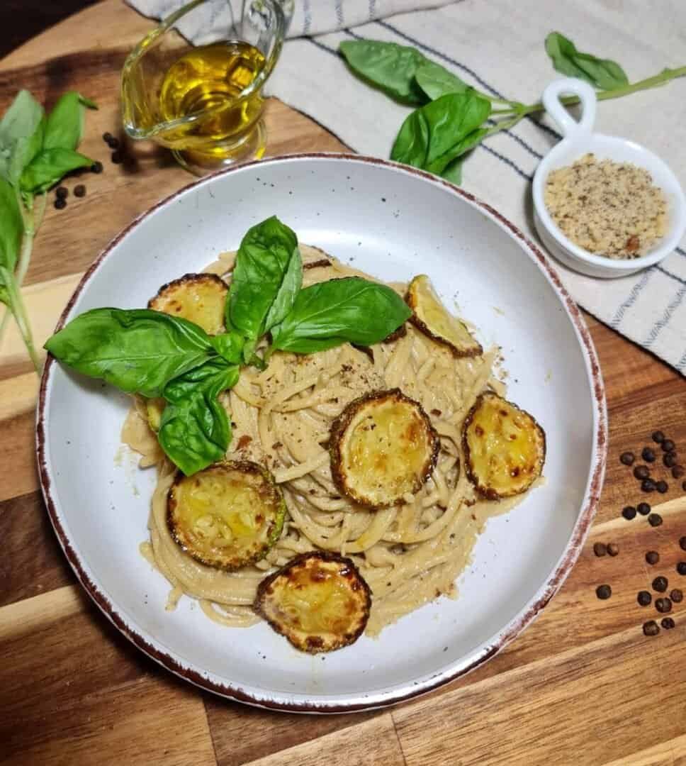 A plate of vegan spaghetti alla Nerano garnished with fresh basil leaves and topped with crispy zucchini slices, surrounded by ingredients including olive oil, ground nuts, and fresh basil on a wooden board.