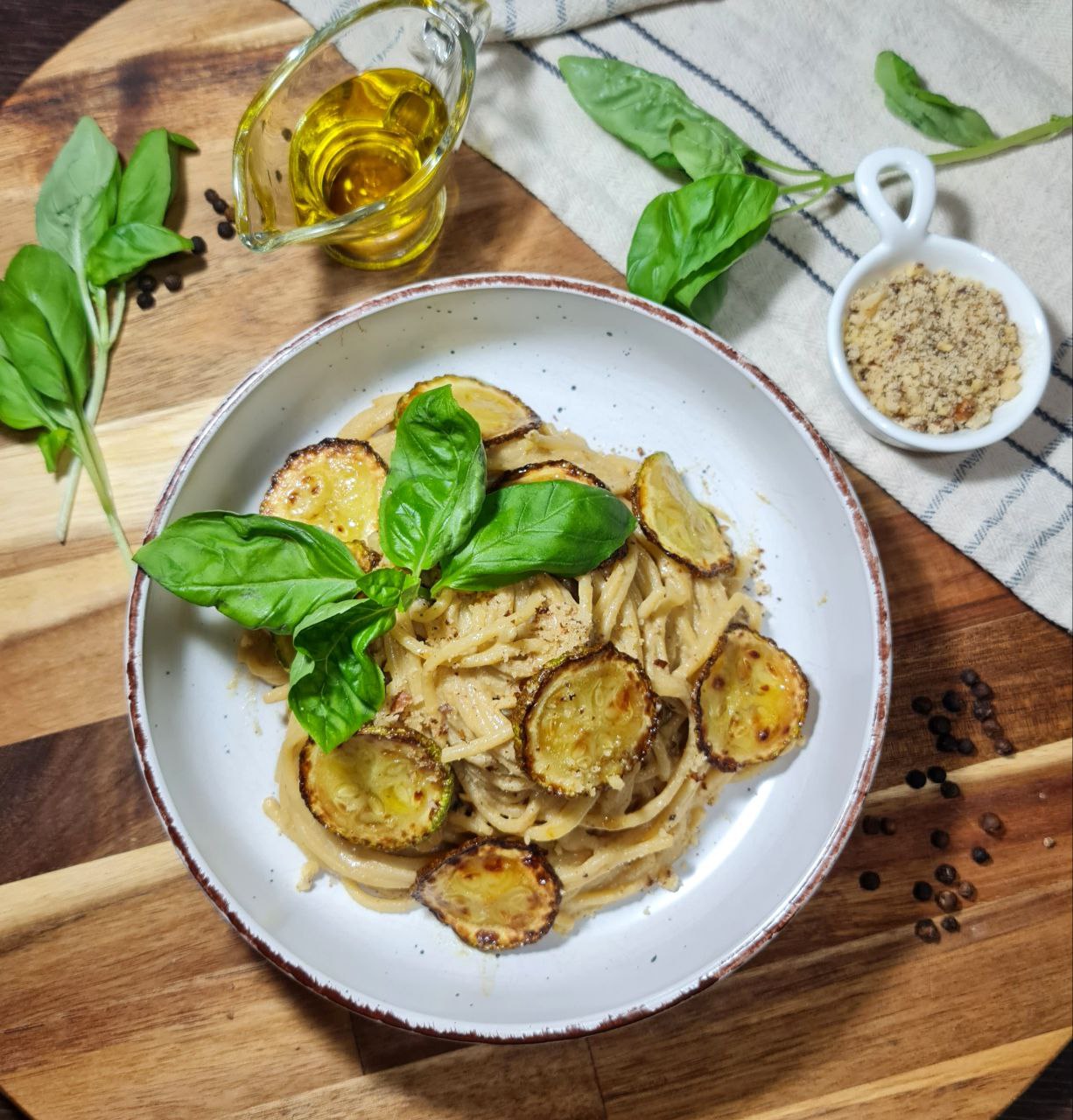 A bowl of vegan spaghetti alla Nerano garnished with fresh basil leaves and roasted zucchini slices, surrounded by ingredients including olive oil, ground nuts, and fresh basil on a wooden surface.